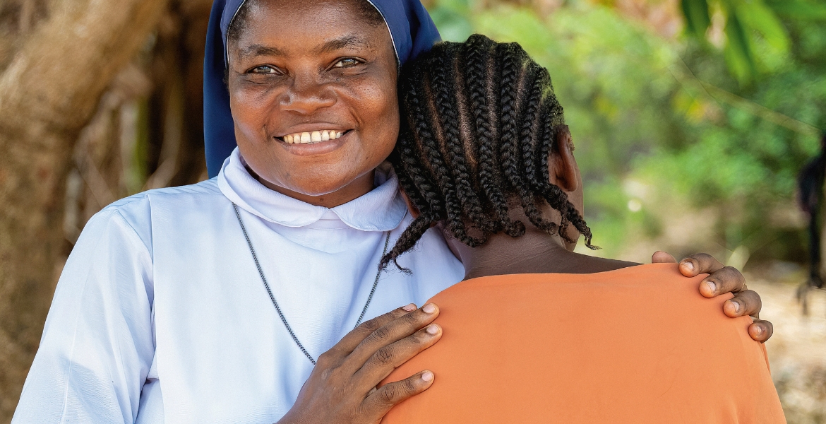 Afrikatag 2027: Eine Ordensschwester mit blauem Schleier und weißem Gewand aus Sierra Leone legt schützend ihre Hände auf die Schultern einer Frau in orangefarbener Kleidung, die dem Betrachter den Rücken zuwendet und ihre Wange an die linke Schulter der Ordensfrau lehnt.
