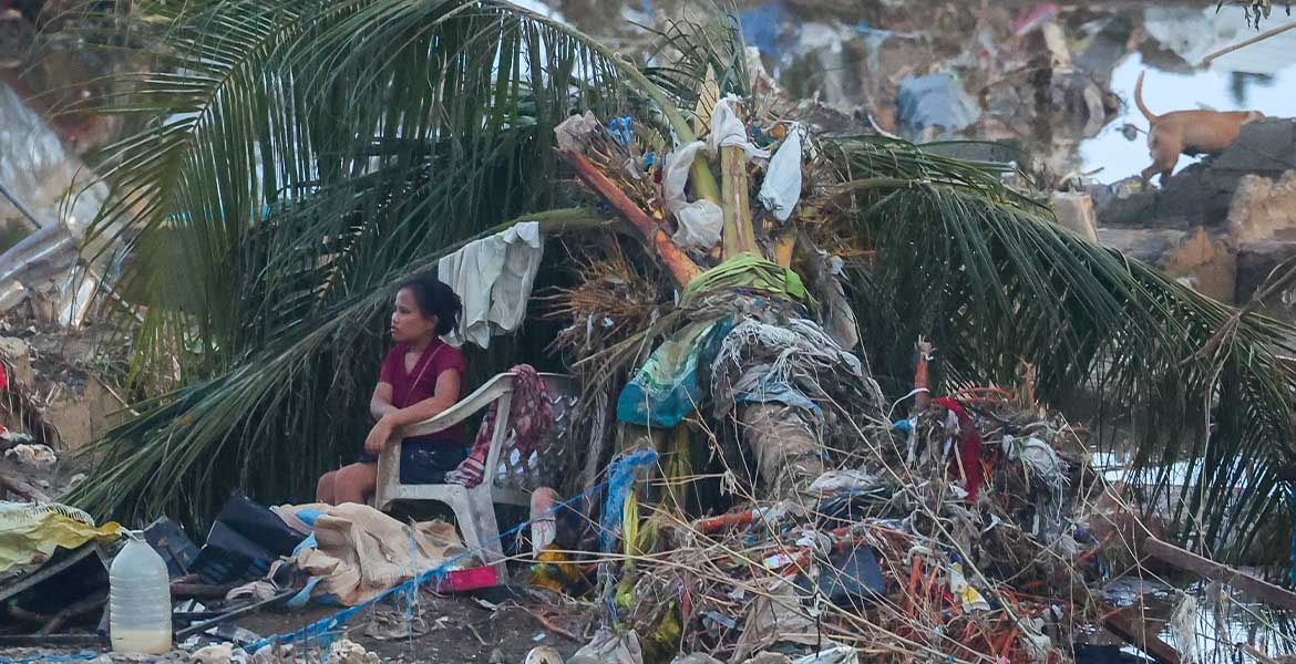 Nach dem verheerenden Taifun Kalmaegi auf der philippinischen Insel Cebu sitzt eine Frau in einem Plastikstuhl unter den Blättern einer umgestürzten Palme. Um sie herum Trümmer, Geröll, Kleiderfetzen und Chaos.