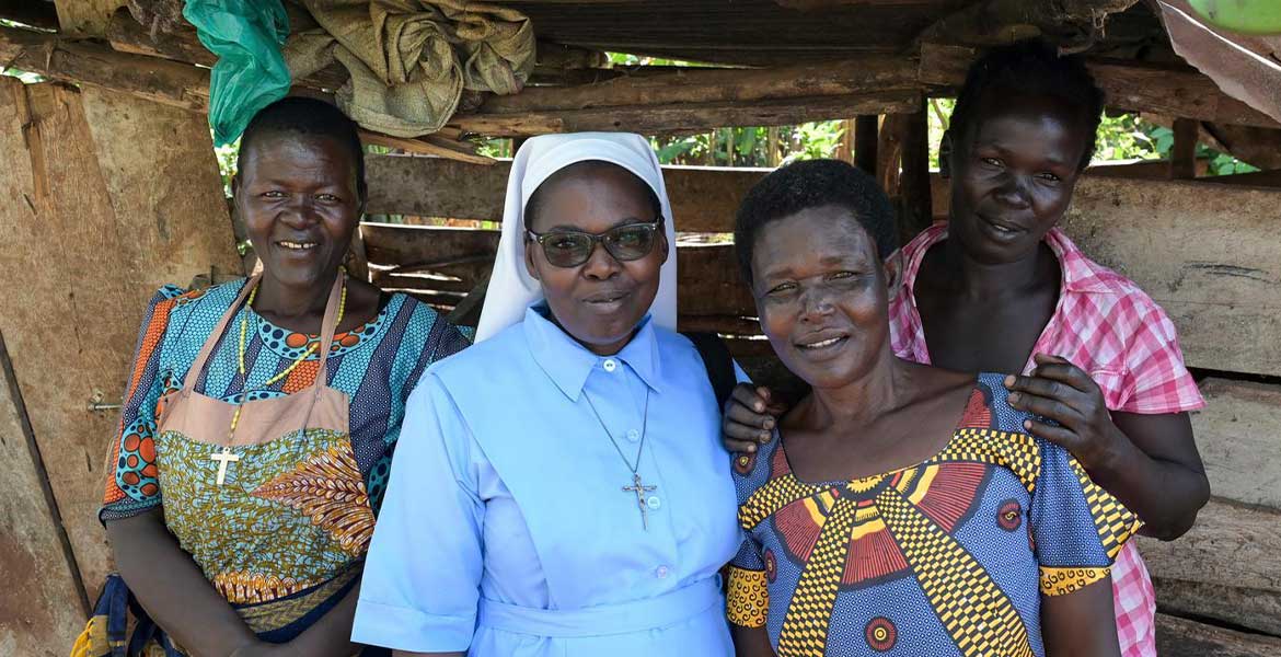 Vier Frauen aus Uganda stehen unter vor einer Bretterwand mit Dach und lächeln in die Kamera. Die zweite von links ist eine Ordensschwester.