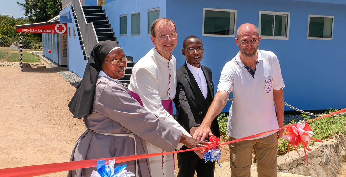 Bei der Einweihung einer Klinik in Tansania durchschneiden missio-Präsident Monsignore Huber gemeinsam mit einer Ordensschwester, einem Priester und dem Missionsarzt Thomas Brei ein rotes Band vor einem blauen Klinikgebäude.