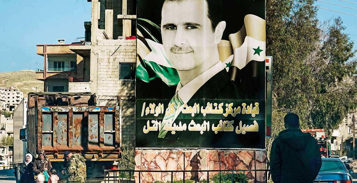 Großes Plakat von Assad an einer Straßenecke. IM Hintergrund Häuser, im Vordergrund ein Mann von hinten.
