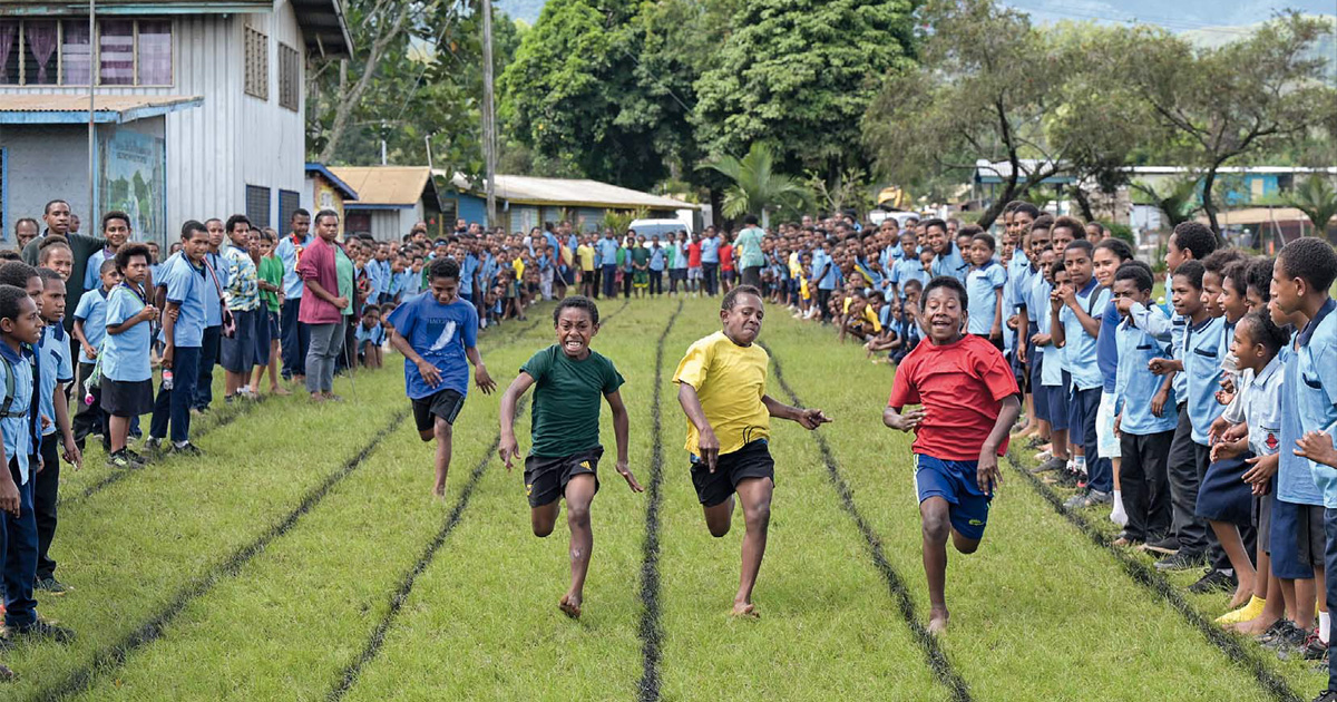 Vier Jungs sprinten um die Wette. Die Laufbahnen sind auf die Wiese gesprüht. Rechts und links davon feuern eine Vielzahl von Kindern und Jugendlichen in Schuluniform die Läufer an.