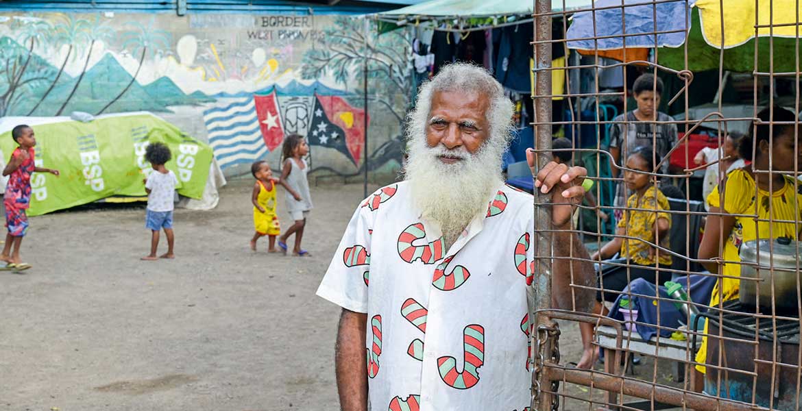 Ein Mann mit grauen Haaren und Bart steht am Tor eines Geflüchteten-Camps. Hinter ihm sind einige Kinder zu sehen. An einer Wand am Ende des Hofs sind zwei Flaggen gemalt, dahinter eine Inselwelt.