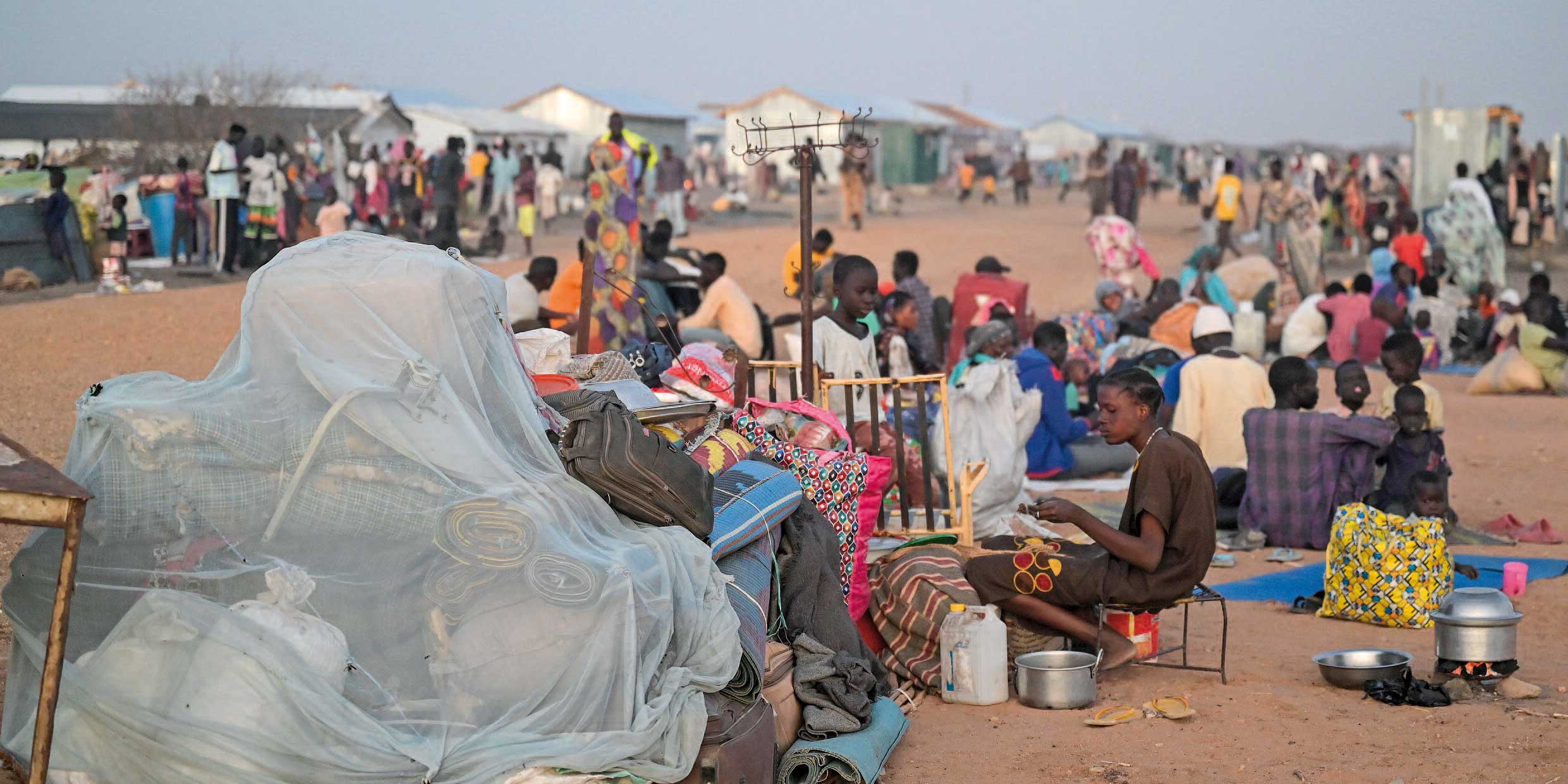 Viele Menschen sitzen unter freiem Himmel in der Abenddämmerung auf einer Straße im Camp, neben sich ihr Hab und Gut.