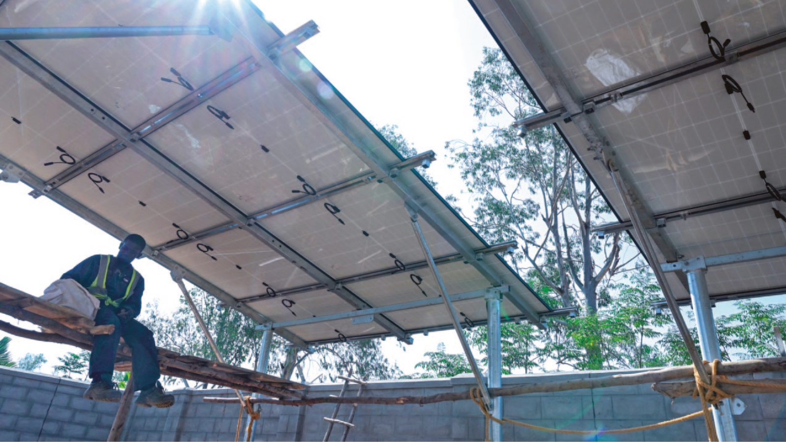 Die Sonne scheint zwischen den zwei Teilen eines Solardachs in Uganda hindurch. Auf dem einfachen Gerüst darunter sitzt ein Arbeiter und macht Pause.