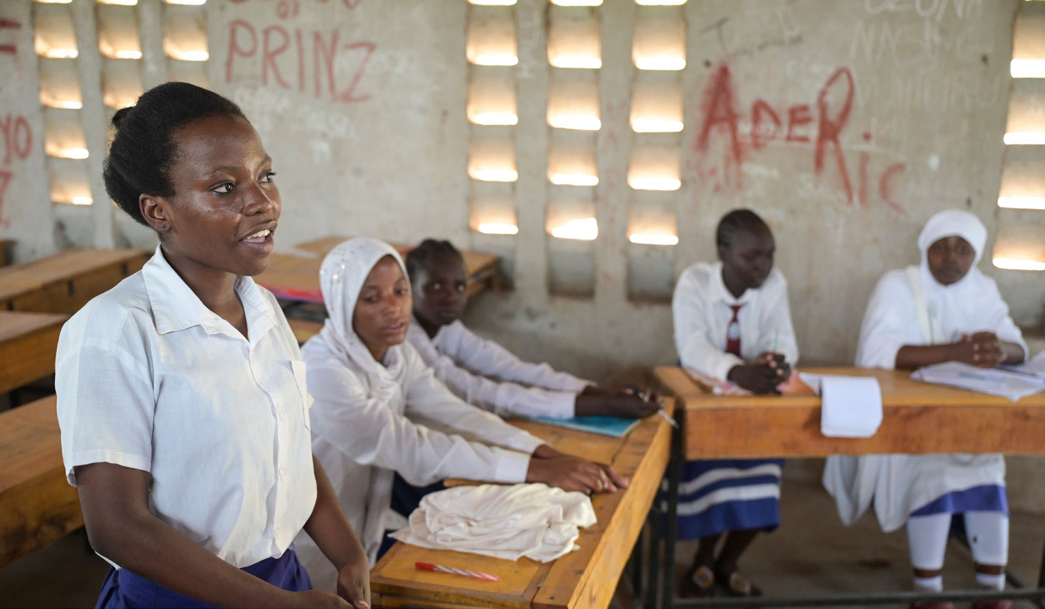 Mädchen steht in einem Klassenzimmer im Geflüchtetencamp Kakuma im Norden Kenias. Nur die erste Reihe ist besetzt. Die übrigen Mädchen sitzen.