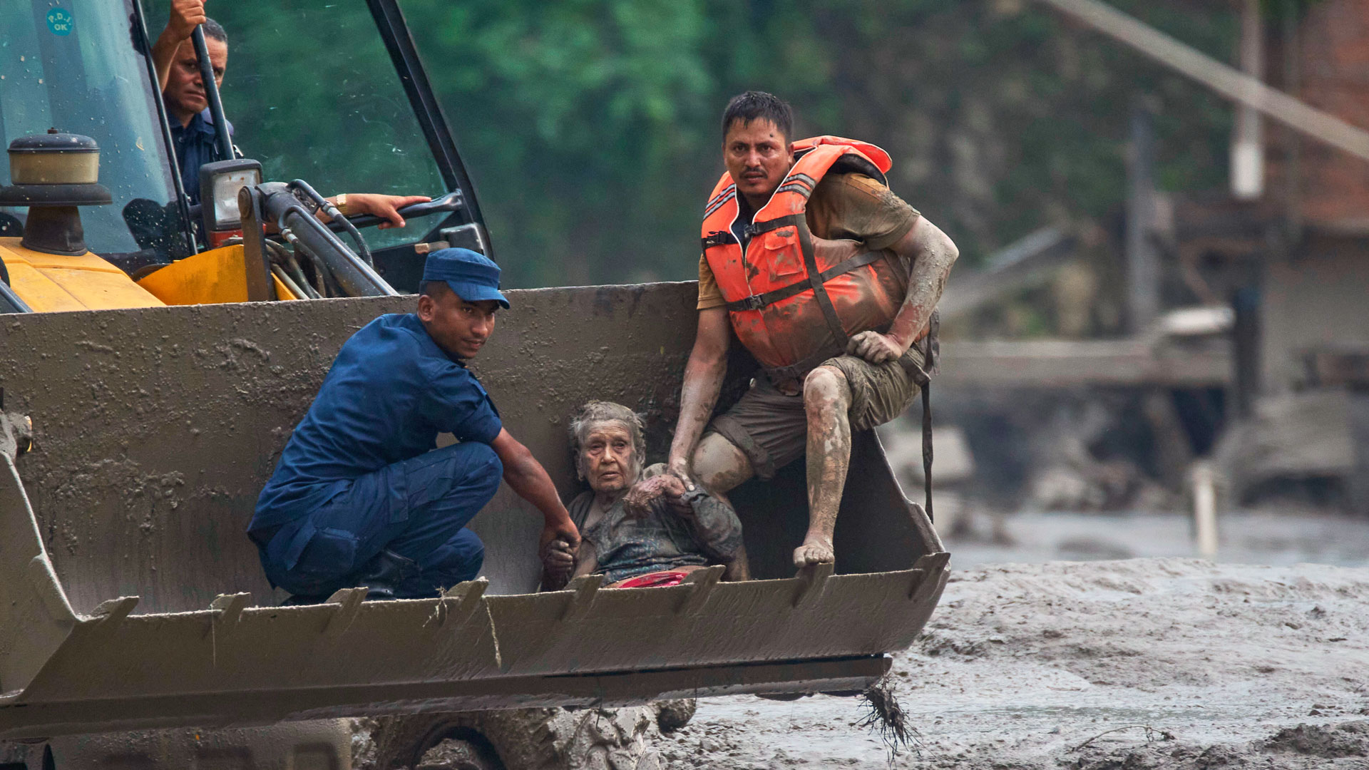 Eine alte Frau sitzt erschöpft mit zwei Helfern in einer Baggerschaufel. Sie wurde zuvor aus den Fluten in Nepal gerettet.