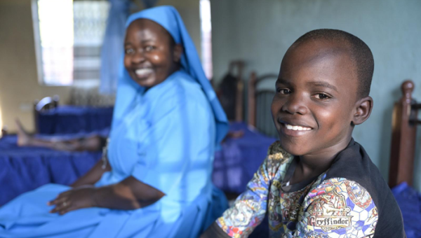 Ein Junge sitzt auf einem Bett im Kinderschutzzentrum Lusubilo in Karonga/Malawi und lacht in die Kamera. Im Hintergrund sitzt eine Ordensschwester und blickt ebenfalls lächelnd in die Kamera.