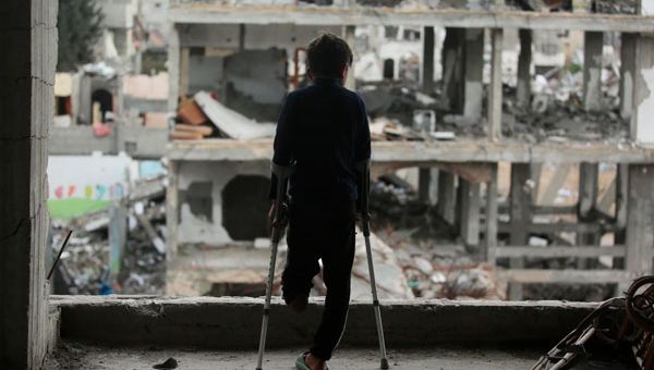 Ein Junge mit amputiertem linken Bein steht auf Krücken und blickt auf Ruinen in Gaza