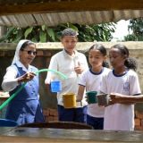 Schwester Rolandine steht mit zwei Schülerinnen und einem Schüler hinter einem Brunnen in einer Schule in Analavory in Madagaskar und gießt mit einem Schlauch frisches Wasser in die Becher der Kinder.
