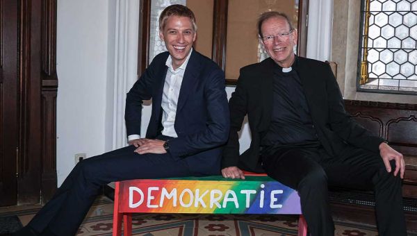 Münchens Oberbürgermeister Dominik Krause (links) sitzt mit missio-Präsident Huber auf einer Bank mit der Aufschrift "Demokratie".".