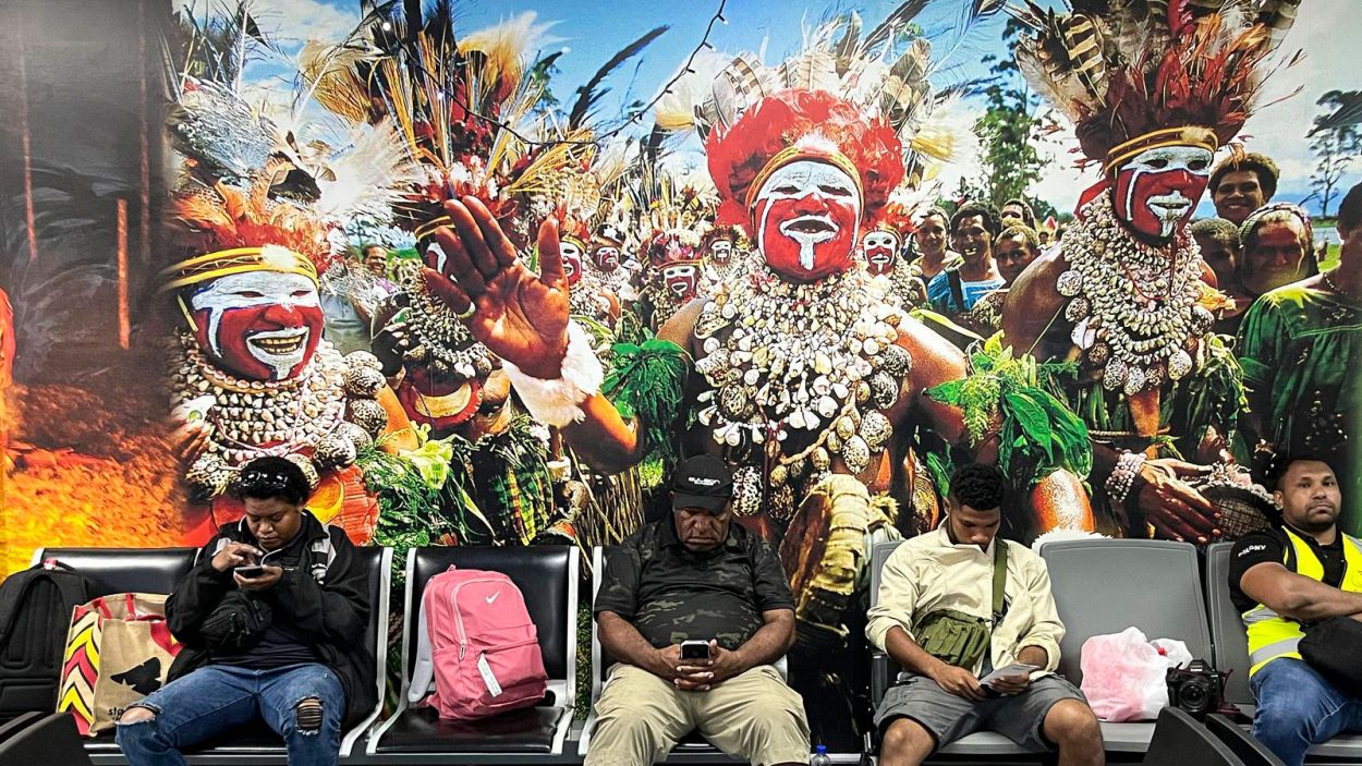 Wartende Passagiere am Flughafen in Port Moresby vor einem Bild von einem Festival in PNG
