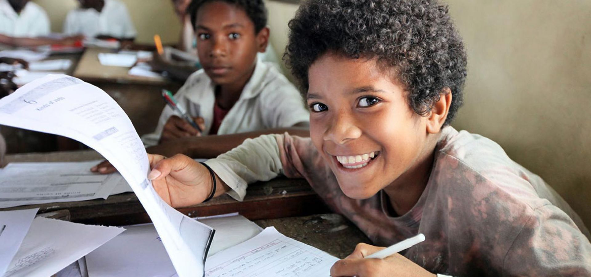 Ein Kind aus Papua-Neuguinea sitzt in der Schule und blickt lachend von seinen Schulunterlagen auf.